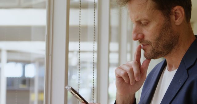 Mature man thoughtfully looking at smartphone while standing by a large window. Ideal for use in technology, business, or lifestyle contexts, conveying themes of concentration, contemplation, and modern communication.