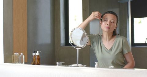 Asian woman applying facial serum at bathroom vanity with round mirror, minimalist decor
