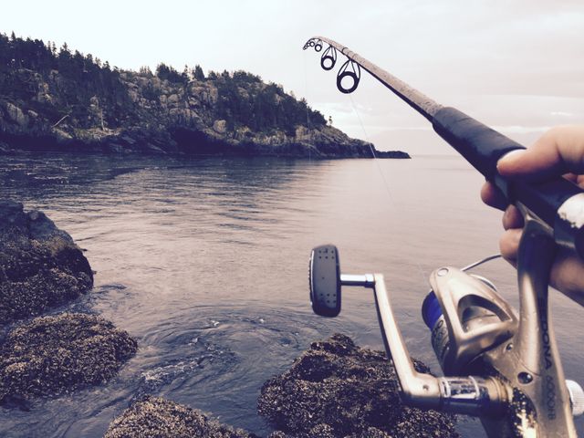 Hand holding fishing rod over rocky coastline with ocean waves, suitable for outdoor adventure, fishing sport promotions, nature-themed content, and travel blogs focused on fishing activities.