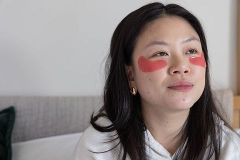 Asian woman wearing red hydrogel under-eye patches relaxing on bed for self-care