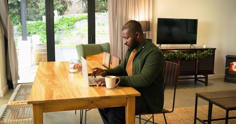 African American man working remotely on laptop at cozy sunlit dining table with coffee
