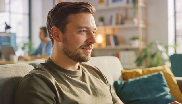 Relaxing bearded man wearing olive tee on sofa in living room with cozy pillows