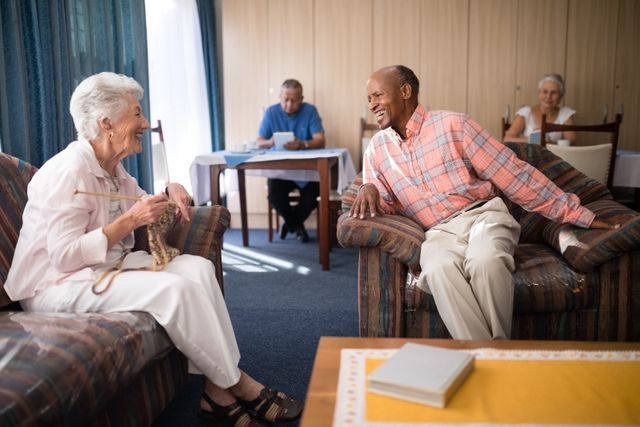 Senior man and woman enjoying a conversation in a comfortable living room at a retirement home. The woman is knitting while the man is smiling and engaging in a friendly chat. This image can be used for promoting senior living communities, retirement homes, elderly care services, and social activities for seniors.
