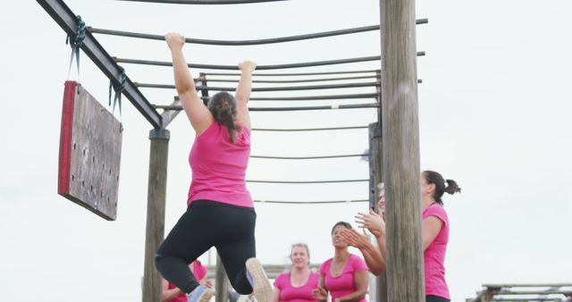 Caucasian female friends in pink t shirts hanging from monkey bars and cheering at bootcamp training. Female fitness, friendship, challenge and healthy lifestyle.