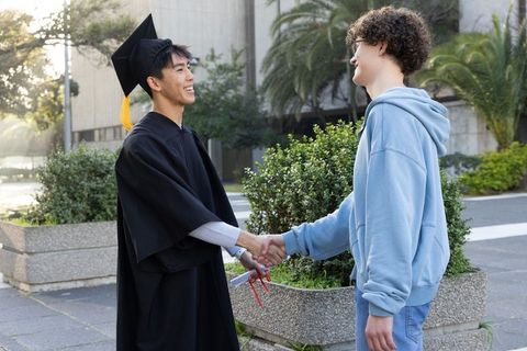 Joyful graduate shaking hands with friend on campus in cap and gown celebrating diploma
