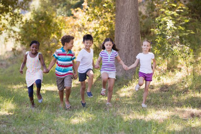 Children are running and holding hands in a grassy outdoor area with trees in the background. They are diverse and appear to be enjoying a summer day, showcasing friendship and playful activity. This image can be used for themes related to childhood, friendship, outdoor activities, summer camps, and diversity.