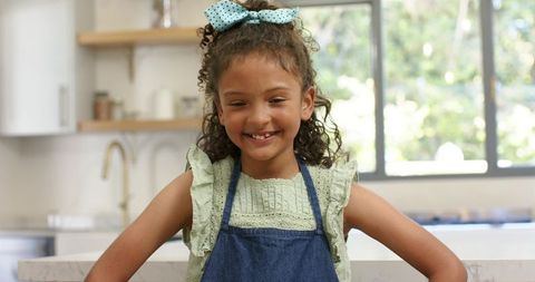 African american girl smiling standing at modern kitchen counter wearing denim apron and bow