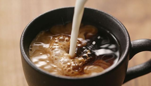 Pouring milk into black ceramic mug of hot coffee creating swirling creamy foam