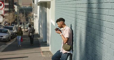 African american man leaning on blue wall eating pastry with headphones in sunlit urban street