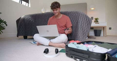 Young man booking travel online while packing carry-on and holding payment card