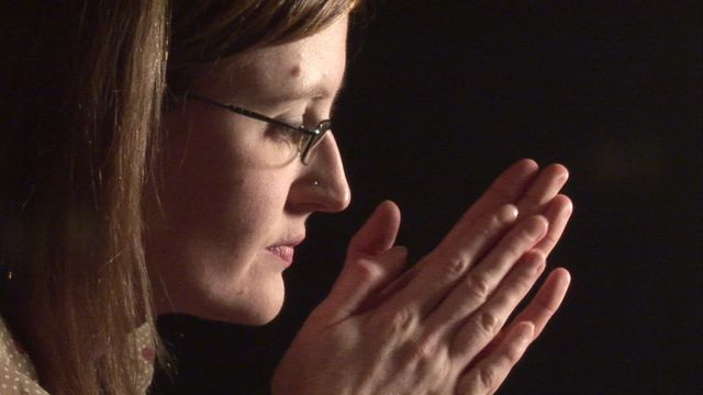 Woman with glasses praying with hands clasped in dim light, highlighting moments of personal reflection and spirituality. Suitable for concepts of faith, contemplation, prayer, hope, devotion, and peaceful solitude. Ideal for use in religious publications, inspirational materials, or projects involving themes of inner strength and resilience.