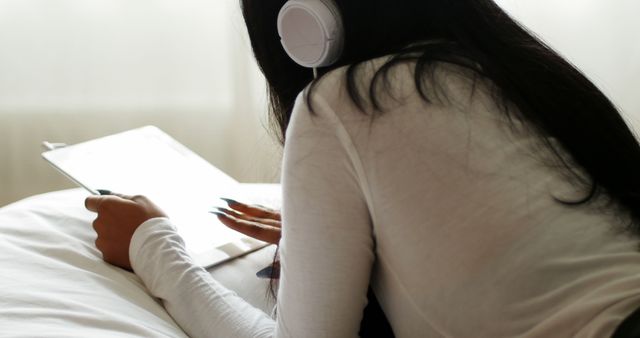 Young woman lying on bed using a tablet while wearing headphones. Ideal for content related to leisure, home comfort, digital technology, and lifestyle. Perfect for articles on relaxation, modern technology usage, and home-based entertainment.
