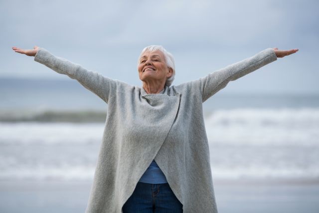 Senior woman standing on the beach with arms outstretched, expressing joy and freedom. Ideal for use in advertisements promoting active lifestyles, retirement planning, health and wellness, or travel. Can also be used in articles about aging gracefully, mental health, and enjoying nature.