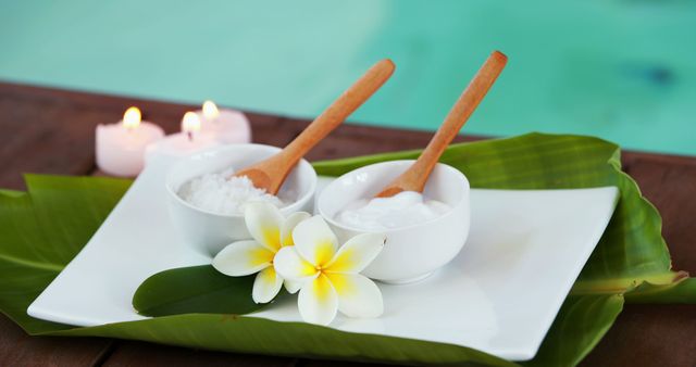 White bowls of spa salt and cream with wooden spoons on large green leaves arranged on tray near pool. Plumeria flowers and burning candles add tranquil and luxurious atmosphere. Ideal for illustrating concepts of spa treatments, relaxation, wellness retreats, and tropical therapies.
