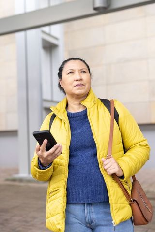 Mature woman wearing yellow coat holding smartphone and looking up on urban sidewalk