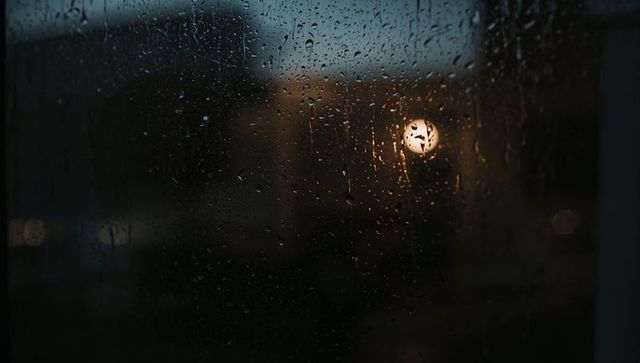 Rainy window showing warm lamp bokeh and urban night reflections, moody interior view