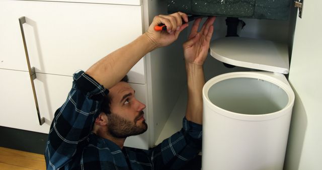 Man repairing plumbing under kitchen sink with tools. Ideal for home maintenance, DIY projects, plumbing services advertisements, handyman guides, or tutorial materials.