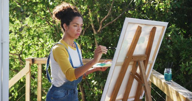 Woman painting on an easel on a wooden deck in a natural outdoor setting with green foliage. Ideal for use in articles or advertisements related to creativity, artistic hobbies, outdoor activities, relaxation, and inspiration. Highlights passion for art and dedication to creative pursuits.