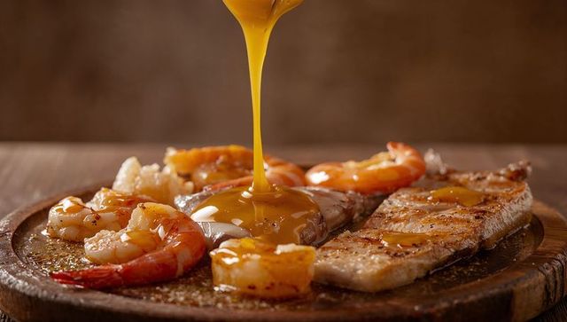 Pouring golden butter sauce over grilled steak and shrimp on rustic wooden platter