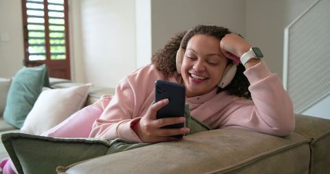 African american woman reclining on sofa listening with headphones, smiling at smartphone