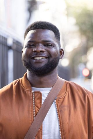 African american man smiling in city wearing rust bomber jacket and crossbody strap, casual
