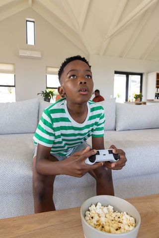 Excited african american boy playing console game on couch with popcorn in living room