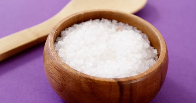 Sea salt is shown in a wooden bowl with a spoon beside it, set against a purple background. Ideal for use in culinary and food-related topics such as cooking, seasoning, health benefits of sea salt, recipe blogs, and kitchen decoration ideas.
