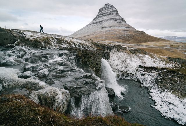 Depicts a lone traveler walking near the iconic Kirkjufell mountain and Kirkjufellsfoss waterfall in Iceland's serene winter landscape. Suitable for travel and adventure articles, nature photography showcases, and tourism brochures focusing on Iceland. Highlights the beauty and allure of Iceland's rugged natural environments.