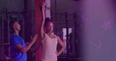 Woman lifting kettlebell overhead in gym with trainer assistance