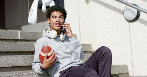 African american student athlete sitting on steps holding football and talking on phone
