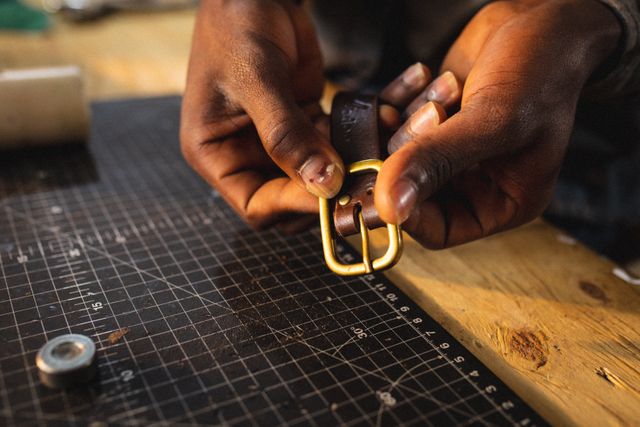 Cropped hands of african american young craftsman showing buckle on leather belt over cutting mat. unaltered, small business, craftsmanship, handcraft, leather craft and workshop.