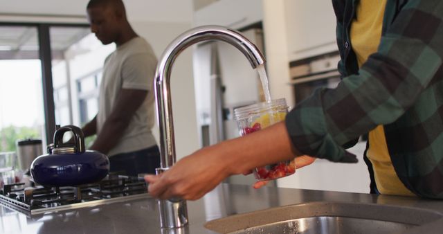 This image depicts two African American individuals preparing drinks in a sleek, modern kitchen. One person is filling a cup with water at the faucet, while the other is working near the stove. The open, bright setting suggests a contemporary home environment. This can be used in articles or advertisements about modern living, home design, kitchen appliances, or healthy habits.