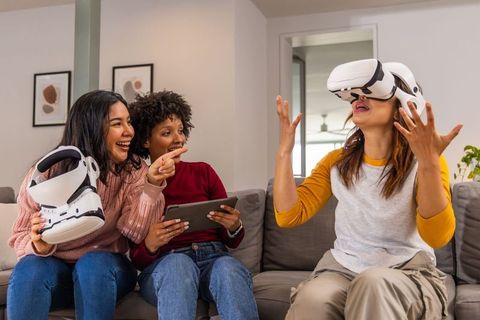 Diverse women testing vr headsets, laughing and pointing on living room sofa