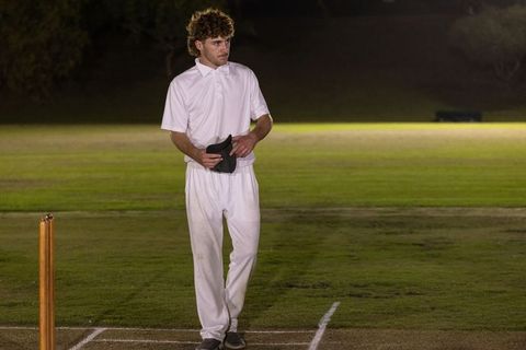 Night cricketer standing by wooden stumps on floodlit grass pitch holding cap before play
