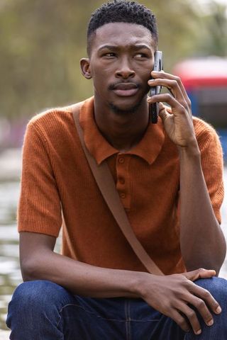 Young man talking on phone by canal wearing rust polo and crossbody bag, urban lifestyle