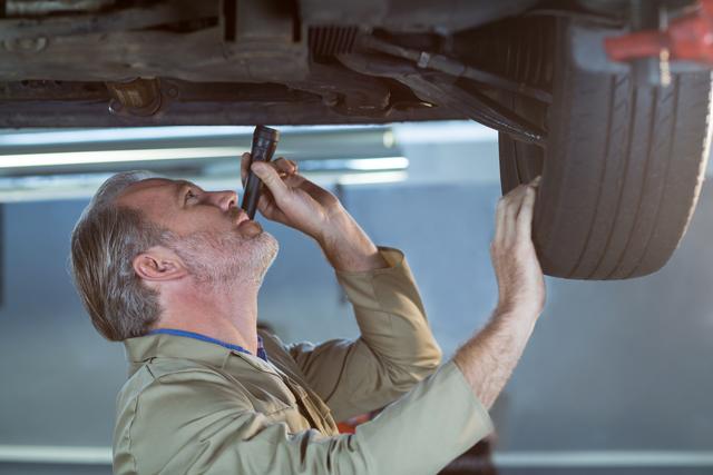 Mechanic inspecting car's undercarriage with flashlight in a repair shop. Ideal for illustrating automotive repair services, professional mechanics, vehicle maintenance, and technical expertise in car workshops. Useful for websites, brochures, and advertisements related to car repair and maintenance services.