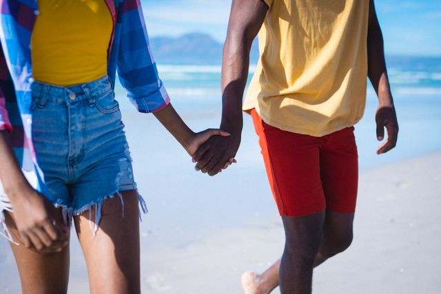 Young African American couple holding hands while walking on a beach during summer. Ideal for use in advertisements, travel brochures, lifestyle blogs, and social media posts promoting love, relationships, and summer vacations.