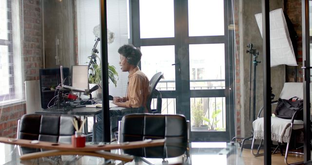 A man is recording a podcast in a modern home office. The scene includes professional setup with a microphone, headphones, desk, and computer. This image is useful for illustrating topics related to podcasting, remote work, technological workspaces, creative jobs, or digital media production.