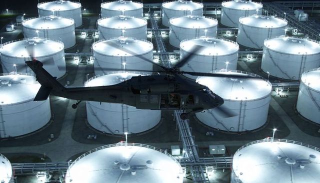 Tactical helicopter hovering above illuminated oil tank farm casting strong rotor shadows