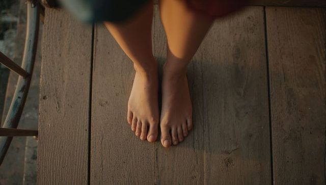 Sunlit bare feet resting on weathered wooden deck with skirt hem and balcony railing at sunset