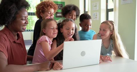 African American teacher guiding diverse elementary students around laptop in classroom