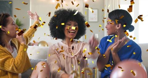 Laughing friends celebrating with gold confetti and balloons on cozy living room sofa