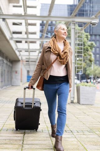Mature woman walking with rolling suitcase through modern urban walkway, travel lifestyle