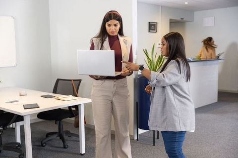 Two professional women collaborating over laptop in modern office workspace