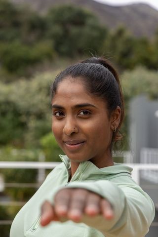Confident indian woman practicing stretching pose on balcony wearing pale green pullover