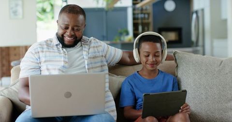 African American father and son using laptop and tablet on couch at home sharing tech time