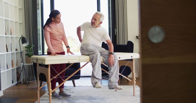 Senior man sitting on a massage table consulting with a physical therapist in a home environment. Man appears to have back pain. Image is useful for topics on elderly healthcare, physiotherapy, in-home medical services, and senior wellness. Ideal for health-related articles, rehabilitation brochures, and caregiver resources.
