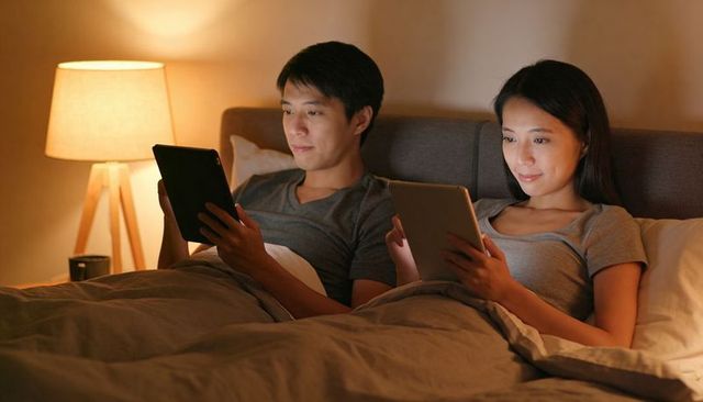Couple reading on tablets in bed at night, relaxing under duvet with warm lamp lighting