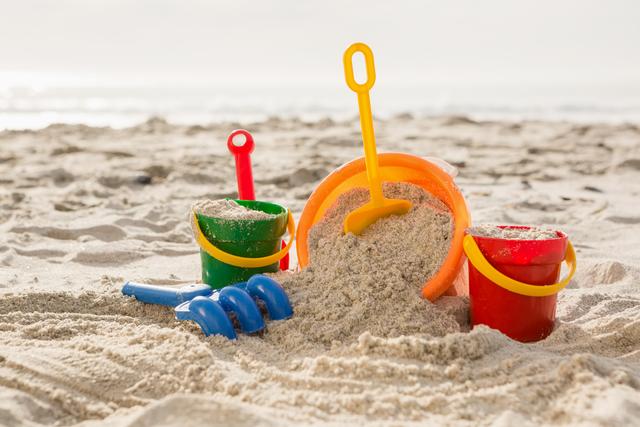 Colorful sand buckets and a shovel are positioned on a sunlit tropical beach, filled with sand and ready for play. Perfect for promoting family vacations, summer activities, relaxation, and childhood memories. Suitable for travel brochures, family blogs, children’s activity guides, and social media posts about beach holidays.