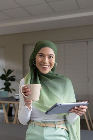 Smiling professional woman wearing green hijab holding mug and tablet in modern workspace